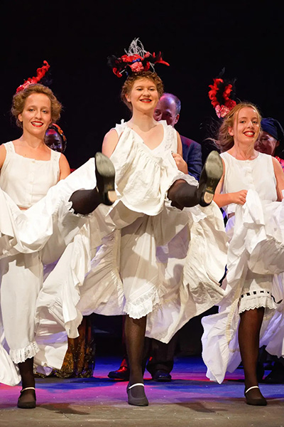 Drei junge Frauen tanzen in historischen Kostümen "Can Can", dahinter mehrere weitere Personen, die applaudieren. Theaterszene aus "Der Kammerdiener" Theatergesellschaft Villmergen